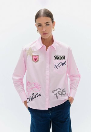 Mujer con una camisa rosa claro abotonada con parches y texto, combinada con vaqueros azul oscuro, de pie con las manos en los bolsillos.