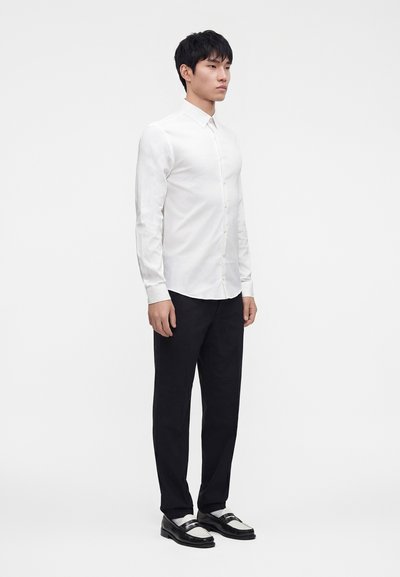 Young man standing wearing a fitted white button-up shirt, black pants, and black and white dress shoes against a plain white background.