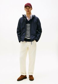 Marine lichtgewicht jack over een wit gestreept sweater, crèmekleurige broek en bruine schoenen. Rode pet maakt de look af met een casual ontwerp.
