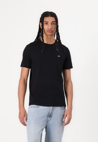 T-shirt noir à manches courtes en coton, avec un col rond et un petit logo blanc sur la poitrine, associé à un jean bleu clair.