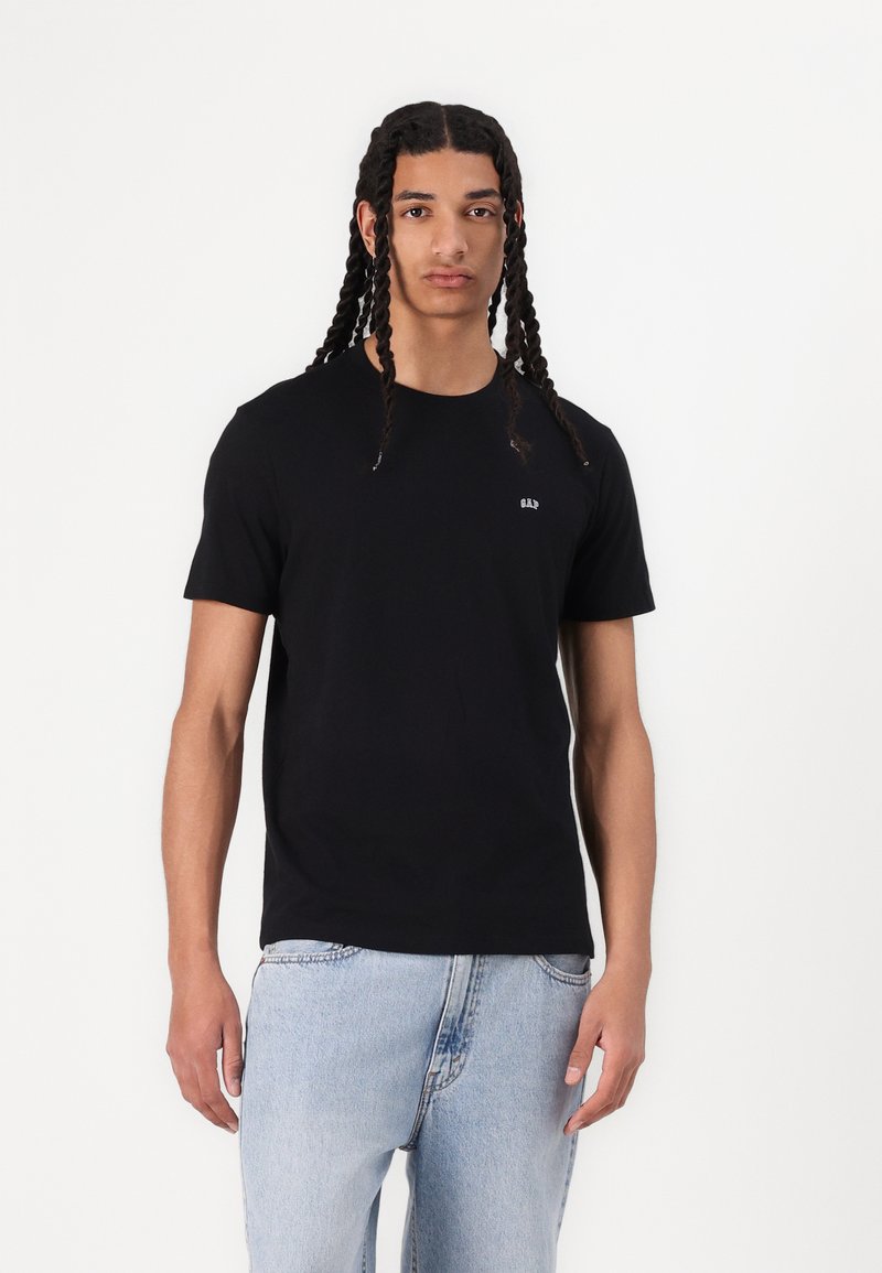 Sort kortærmet t-shirt i bomuld med rund halsudskæring og et lille hvidt logo på brystet, iført med lyseblå jeans.