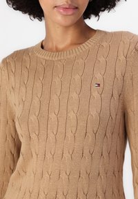 Pull en tricot câble beige avec un col rond et un petit patch logo présentant des accents rouges, blancs et bleus sur la poitrine.