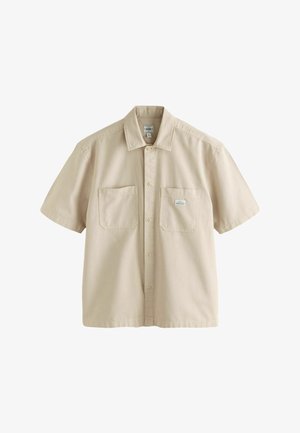 Bež srajca s kratkimi rokavi iz bombaža, ki ima gumbasto zapiranje spredaj, dve prsni žep, in ravno obrobo z diskretnimi šivnimi podrobnostmi.