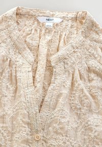 Blouse beige brodée avec des motifs floraux, décolleté en V, tissu texturé et fermeture centrale à boutons. L'étiquette indique "NEXT."