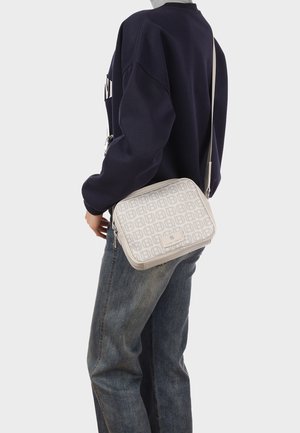 Persona che indossa un maglione scuro e jeans porta una borsa a tracolla bianca con motivo geometrico ripetuto e tracolla regolabile.