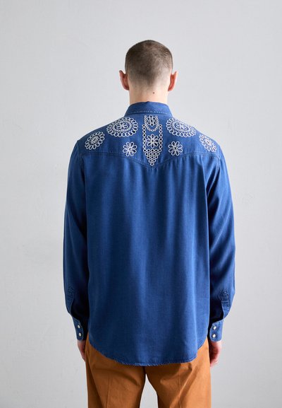 Blaues Denimhemd mit langen Ärmeln, das auf dem Rücken eine weiße bestickte Blumenmusterung und einen abgerundeten Saum aufweist, kombiniert mit braunen Hosen.