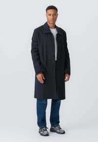 Ung mand står, iført en lang sort frakke, hvid t-shirt, blå jeans og grå sneakers mod en ensfarvet lys baggrund.