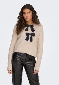 Beige neulottu neuletakki, jossa on pyöreä pääntie ja kaksi mustaa nauhabroodia edessä. Siinä on pitkät hihat ja hieman lyhennetty helma.