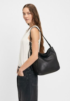 Donna con capelli lunghi e ondulati, che indossa una canotta bianca senza maniche e jeans neri, mentre porta una grande borsa a tracolla in pelle nera.
