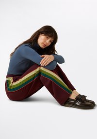 Blå ribbat polotröja, kastanjefärgade byxor med gröna och gula ränder, och bruna fransade loafers. Modell sitter med armarna omkring knäna.