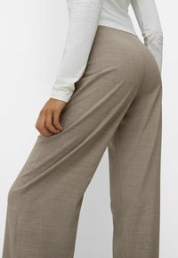 Spodnie o szerokich nogawkach wykonane z jasnobrązowej tkaniny o teksturowanym wykończeniu. Posiadają boczne kieszenie i luźny fason, zestawione z białą bluzką z długim rękawem.