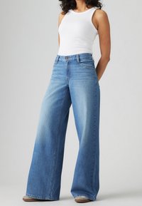 En person som bär en vit ärmlös topp och högmidjade vida jeans i blått, står mot en ljus bakgrund.