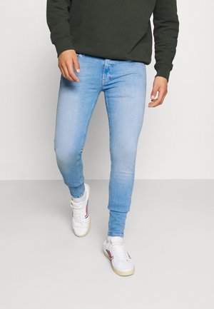 Homme portant un jean skinny bleu clair, un sweat-shirt vert foncé et des baskets blanches marchant sur une surface unie gris clair.