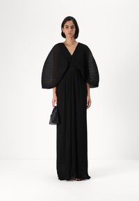 Čierne maxidress s plisovaním, hlbokým V-výstrihom a dramatickými batwing rukávmi. Model drží malú čiernu kabelku a stojí na jednoduchom pozadí.