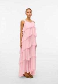 Abito a strati rosa con ruches, realizzato in tessuto leggero. Presenta spalline larghe e scende fino alla caviglia, abbinato a scarpe verde oliva.
