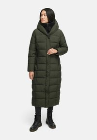 Lång, olivgrön puffjacka med huva, featuring kviltade sektioner, framdragkedja och sidofickor. Bärs över svart outfit och stövlar.