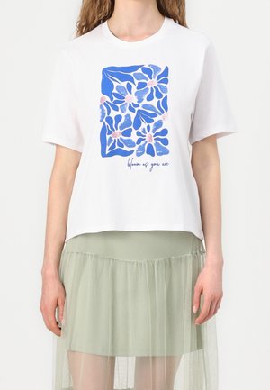 Valkoinen lyhythihainen t-paita, jossa on sininen kukkakuva ja teksti "bloom as you are" edessä, yhdistetty vaaleanvihreään kerrokselliseen tyllihameeseen.