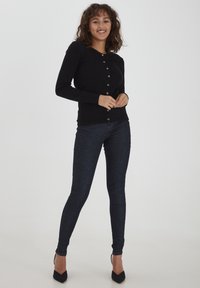 Cardigan nero con bottoni e maniche lunghe, caratterizzato da un design aderente e bottoni argentati, abbinato a jeans skinny in denim scuro e tacchi neri.
