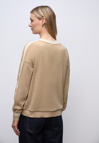 Sudadera de color beige claro con cuello y puños acanalados, que presenta rayas laterales en crema. Textura suave, ajuste relajado, combinada con jeans azul oscuro.
