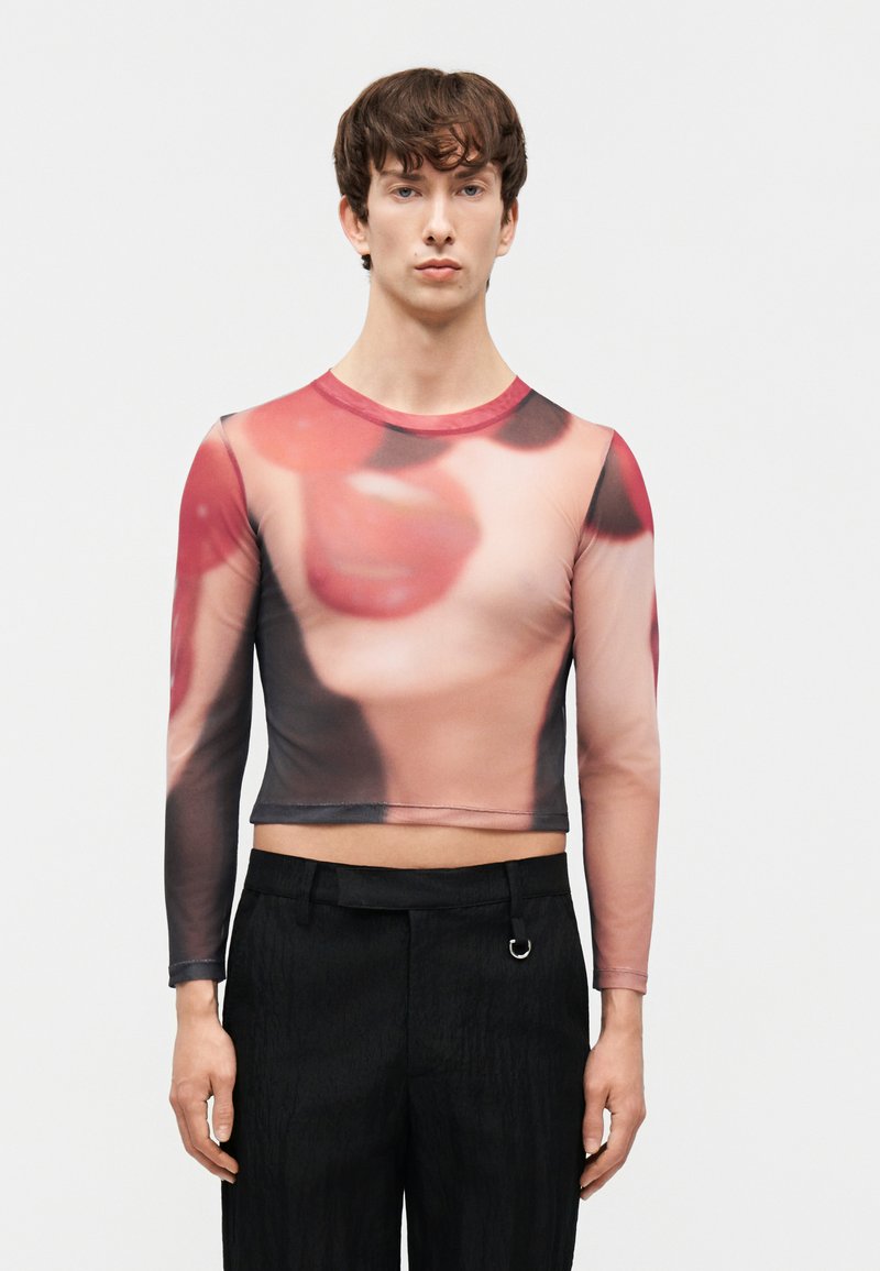 Hombre que lleva una camiseta corta semitransparente de manga larga con una impresión grande y desenfocada en primer plano de unos labios rojos y un rostro en colores suaves.