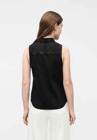 Γυναίκα με καστανά ίσια μαλλιά που φοράει αμάνικο μαύρο πουκάμισο από τζιν και άσπρο παντελόνι, γυρισμένη προς τα πίσω, σε απλό ανοιχ τό φόντο.