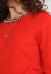 Suéter rojo de punto con cuello redondo y ribete a rayas en contraste, textura suave y ajuste relajado, combinado con un collar sencillo.