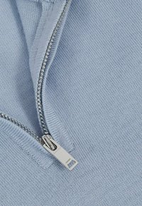 Világoskék kötött anyag, ezüst cipzárral. Közeli nézet, amely kiemeli a cipzár részleteit és az anyag finom textúráját.