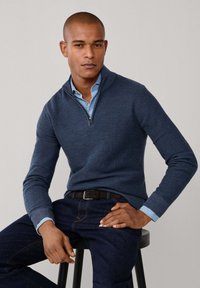 Maglione in maglia navy con zip a metà e polsini a coste, indossato sopra una camicia colletto azzurra, abbinato a jeans in denim scuri e una cintura nera.
