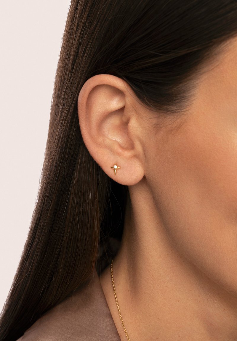 Pendiente de oro en forma de estrella, con una pequeña piedra preciosa clara en el centro, llevado en la oreja. Piel suave y cabello oscuro visible.