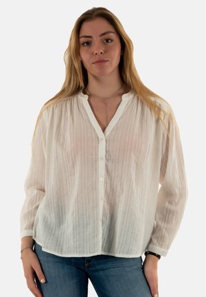 Camisa blanca de manga larga hecha de tejido texturizado. Cuello tipo notch y cierre frontal con botones, con sutiles rayas verticales. Combinada con jeans azules.