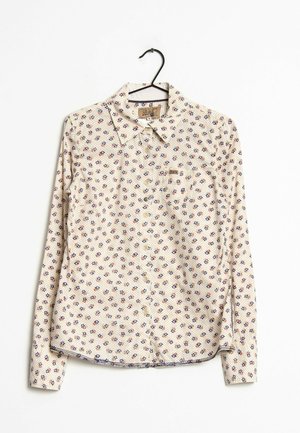 Langærmet beige skjorte med knapper og små røde og blå blomsterprint, hængende på en sort bøjle mod en hvid baggrund.