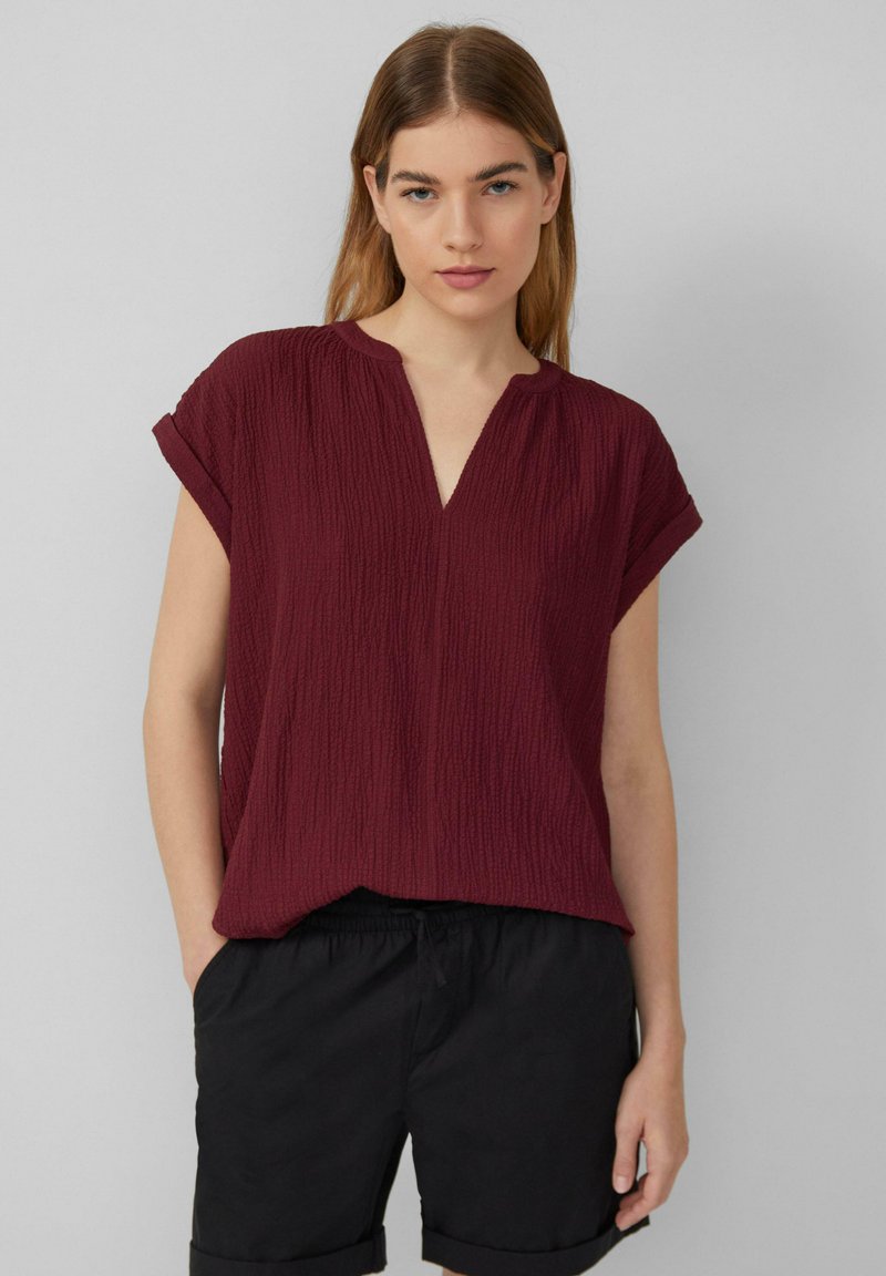 Bordeauxfarbene Kurzarmbluse mit strukturiertem Material, V-Ausschnitt und lockerer Passform. Kombiniert mit schwarzen Shorts mit einem elastischen Bund.