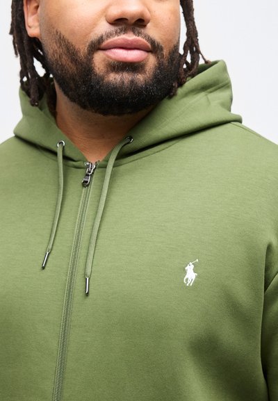 Άνδρας με γενειάδα και κότσους dreadlocks που φοράει πράσινο φούτερ με φερμουάρ και λευκό λογότυπο ποδοσφαιριστή στο στήθος, μπροστά σε λευκό φόντο.