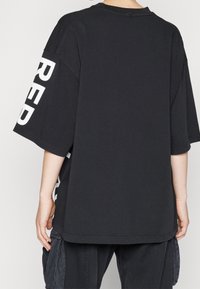 Osoba oblečená v černém oversized tričku s velkým bílým textem na levém rukávu a volných černých cargo kalhotách, stojící na světlém pozadí.