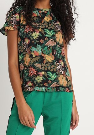 Vrouw draagt een zwart kortärmelig shirt met een kleurrijk bloemen- en dierenprint, gecombineerd met groene broek met zwarte zijkstrepen.