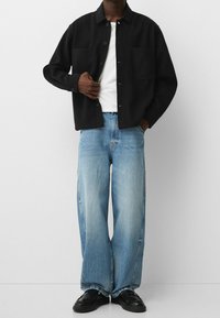 Svart knäppskjorta med två framfickor, kombinerad med löst sittande ljusblå jeans och svarta skor, som framhäver tygtextur och passform.