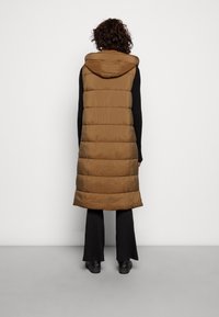 Brązowa ocieplana długa kamizelka z kapturem, z pionowymi przeszyciami i prostym krojem, zestawiona z czarnymi spodniami w rozszerzanym kroju i czarnymi butami.