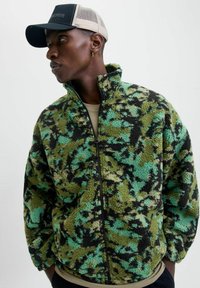 Fleece jack in groen, zwart en turquoise camoprint met een hoge kraag en een rits aan de voorkant. Model draagt een zwarte pet met een mesh achterkant.