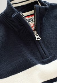 Marineblauer Strickpullover mit weißem Streifen, halb mit silbernem Reißverschluss geschlossen, gerippter Kragen und sichtbarem roten und weißen Markenetikett im Kragen.