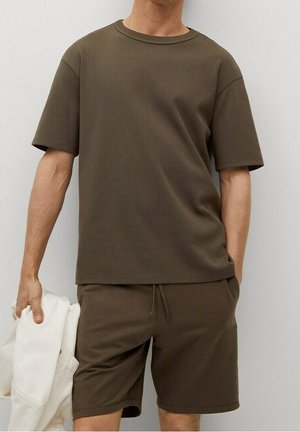 T-shirt marron à manches courtes avec un col rond, assorti à un short. Texture douce et lisse, coupe décontractée. Main tenant une veste crème.