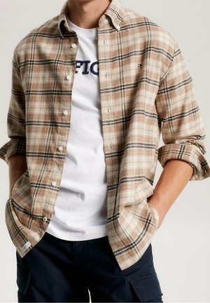 Personne portant une chemise en flanelle à carreaux beige sur un t-shirt graphique blanc et un pantalon foncé, les mains dans les poches.