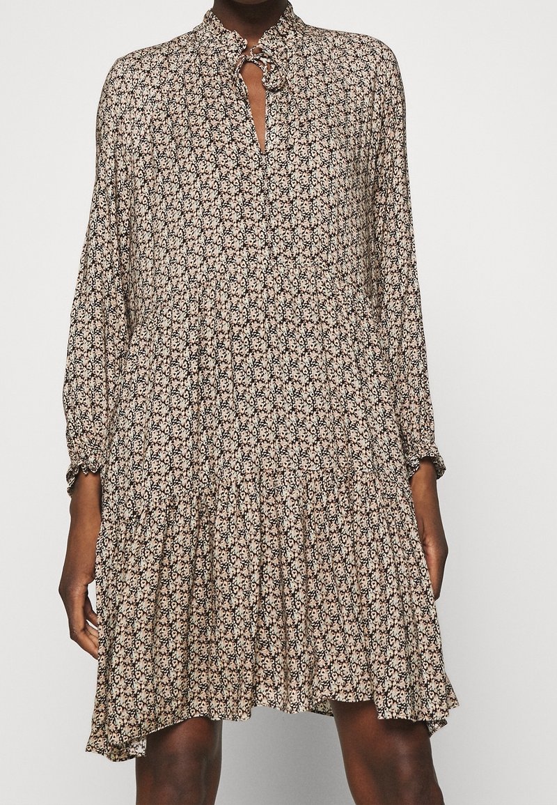 Robe noire et beige à motifs, avec un col arrondi, des manches longues et une jupe à volants. Présente une ouverture en forme de clé avec un accent circulaire.