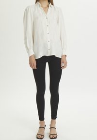 Cremestripenhemdel mit Knopfbesatz, langen Ärmeln und puffigen Schultern, kombiniert mit schwarzen, eng anliegenden Hose und schwarzen, offenen High Heels.