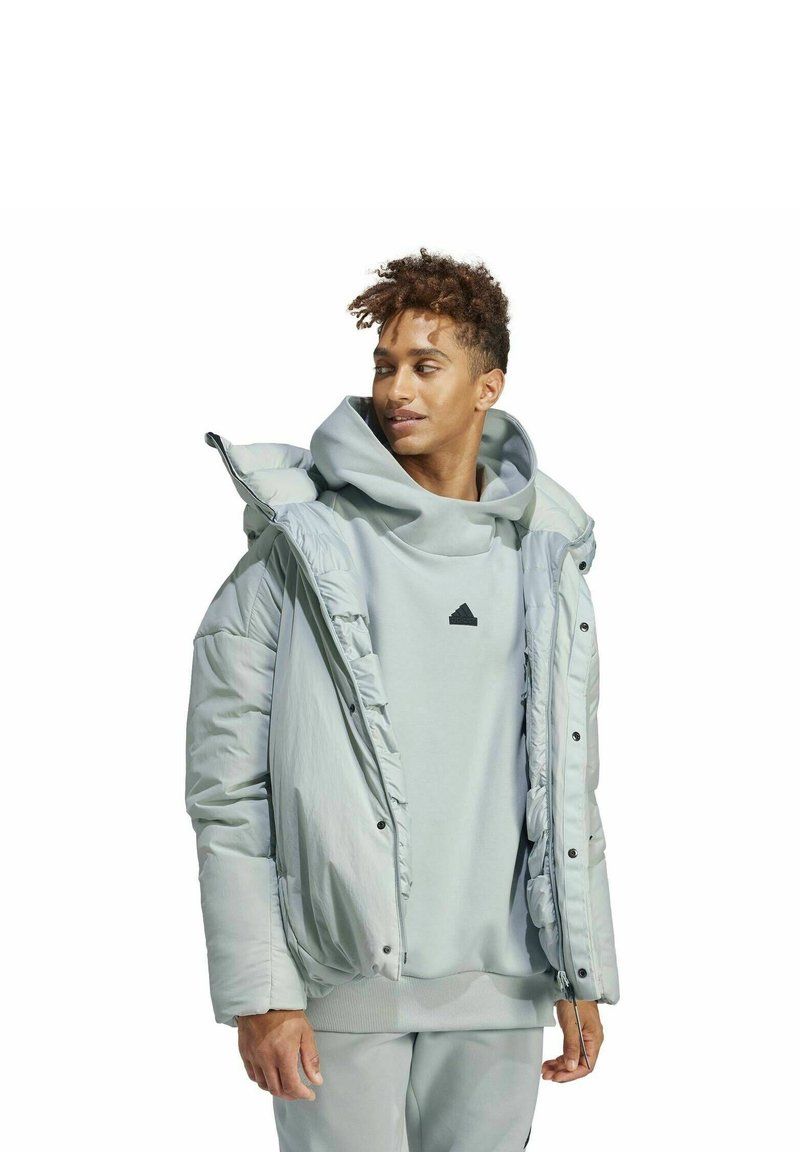 Jasnoszara puchowa kurtka o fakturowym wykończeniu, z kapturem i zamek błyskawiczny z przodu. Nałożona na jasnoszary hoodie z czarnym logo.