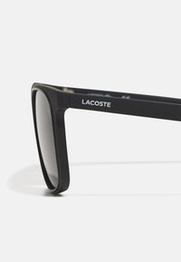 Ochelari de soare negri cu finisaj mat, formă rectangulară și lentile întunecate. Logo-ul Lacoste este afișat în alb pe partea ramei.
