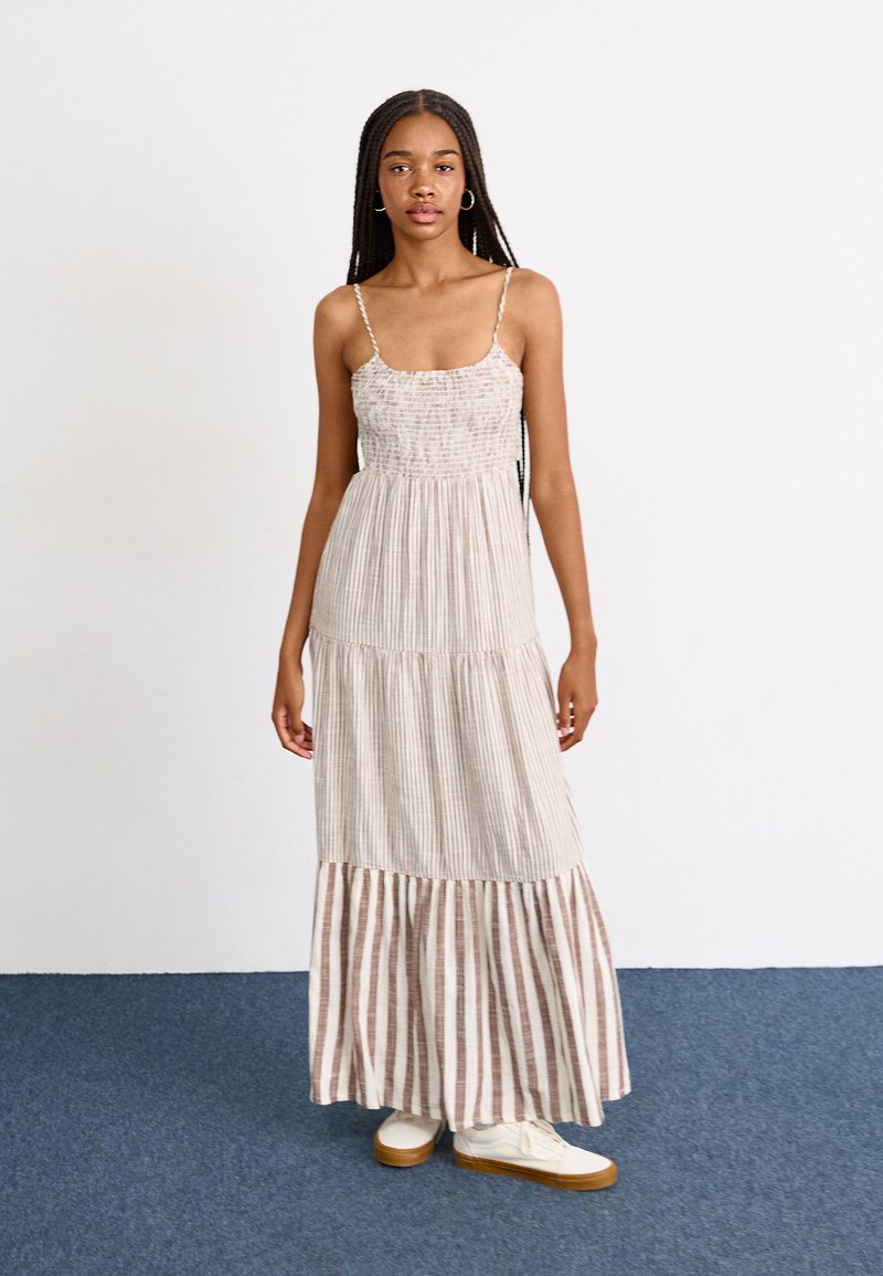 Robe maxi avec des petites bretelles, bodice froncé et tissu rayé en beige et marron. Ourlet évasé en étages et portée avec des baskets blanches.