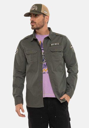 Homme portant une chemise verte boutonnée avec deux poches poitrine et des écussons, un t-shirt graphique violet, un pantalon noir et une casquette verte et beige.