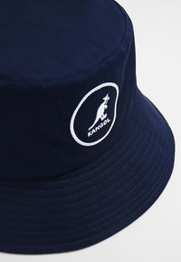 Cappello a secchiello color blu navy realizzato in tessuto con una corona piatta e un ampio bordino. Presenta un logo di canguro bianco all'interno di un design circolare.
