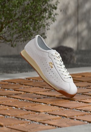 Sneaker in pelle bianca con perforazioni, lacci bianchi, suola beige e logo coccodrillo dorato, posizionata sopra piastrelle di legno all'aperto.