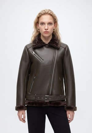 Femme aux cheveux blonds portant une veste en cuir marron foncé avec un col et des poignets en fourrure, debout devant un fond clair uni.